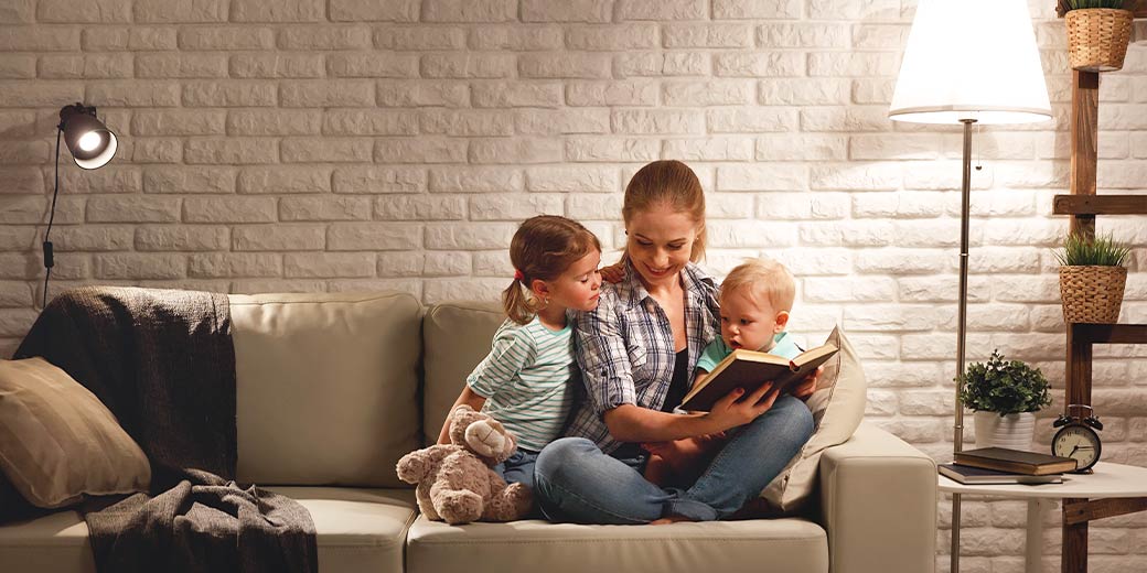 Mutter sitzt eng mit einer Tochter und einem Sohn abends auf dem Sofa und sie lesen gemeinsam ein Buch