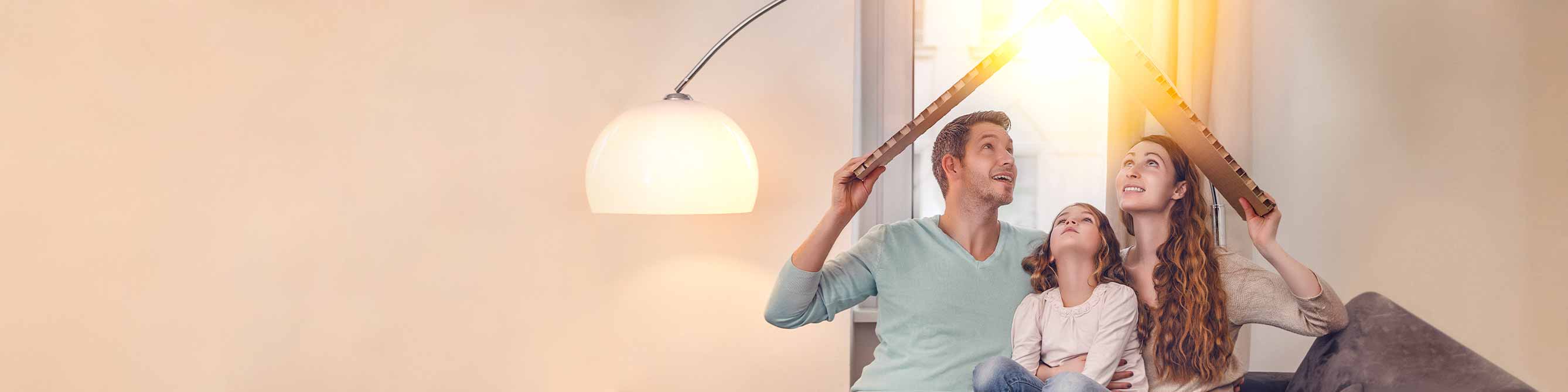 Familie sitzt auf einem Sofa und hält sich ein Dach aus Pappe über die Köpfe